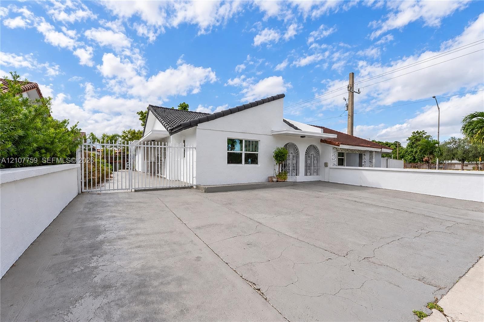 Single-story residence featuring a white stucco exterior, dark tile roof, arched entryway accents, and a gated concrete parking area enclosed by a perimeter wall