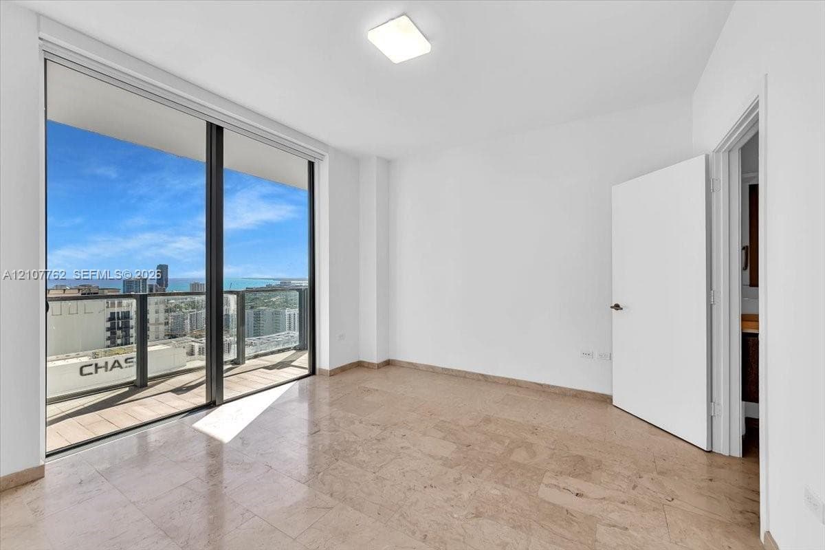 Floor-to-ceiling sliding glass doors, private balcony, neutral stone tile flooring, and overhead flush-mount lighting