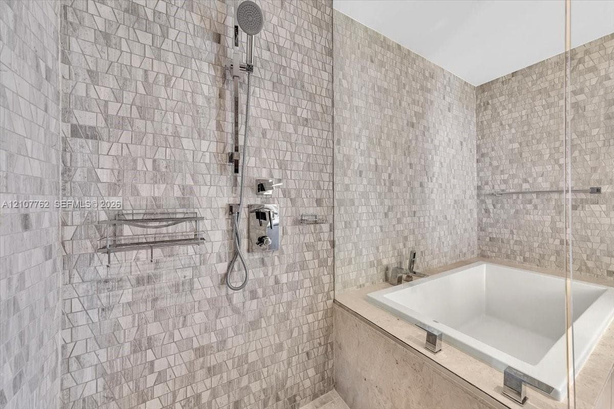 Soaking tub with stone-finish surround, glass shower enclosure, mosaic tile wall detailing, handheld shower fixture, and polished chrome hardware