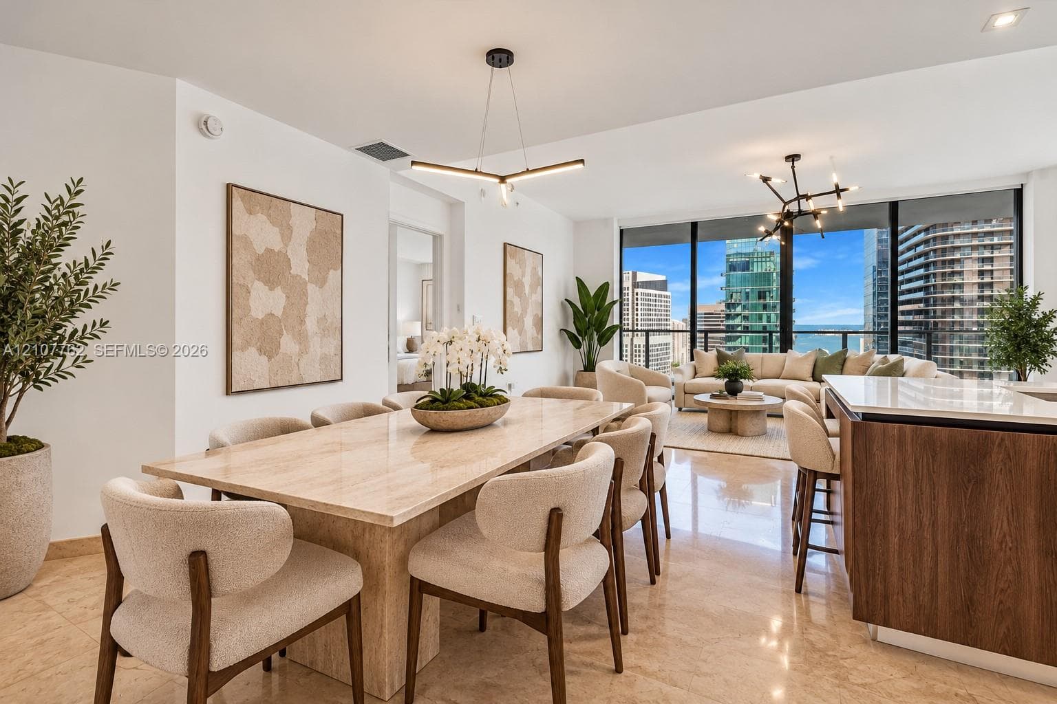 Open-concept interior featuring floor-to-ceiling windows, polished stone flooring, modern linear chandeliers, and an island with a wood-finish base and light-toned stone countertop