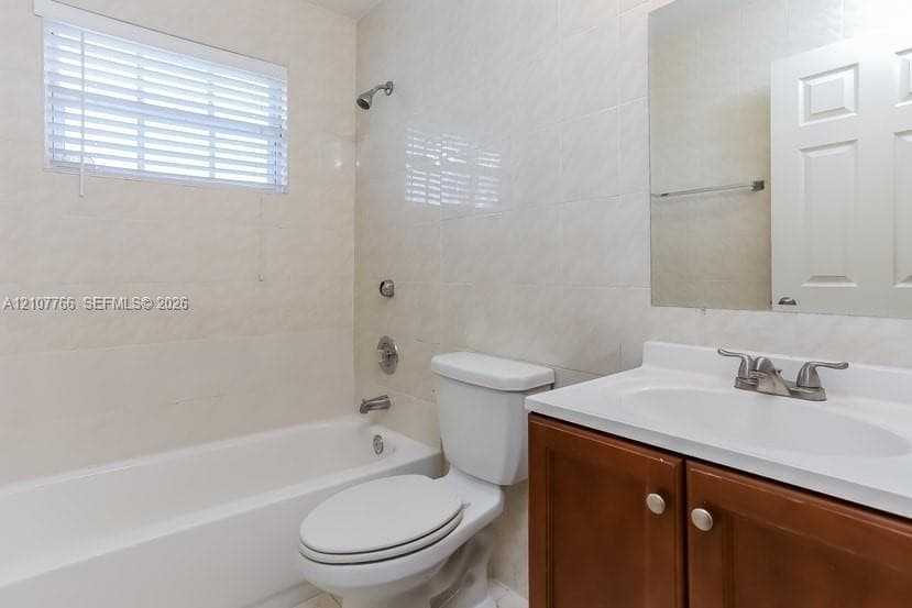 Bathroom featuring full-height wall tiling, integrated tub-shower combination, wooden vanity cabinet, and rectangular wall-mounted mirror