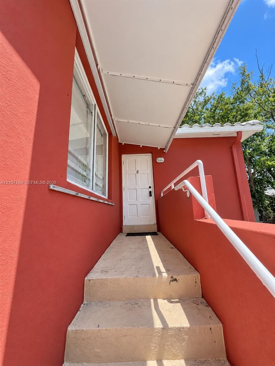 Exterior entrance featuring a white panel door, concrete steps, a metal handrail, and a covered walkway