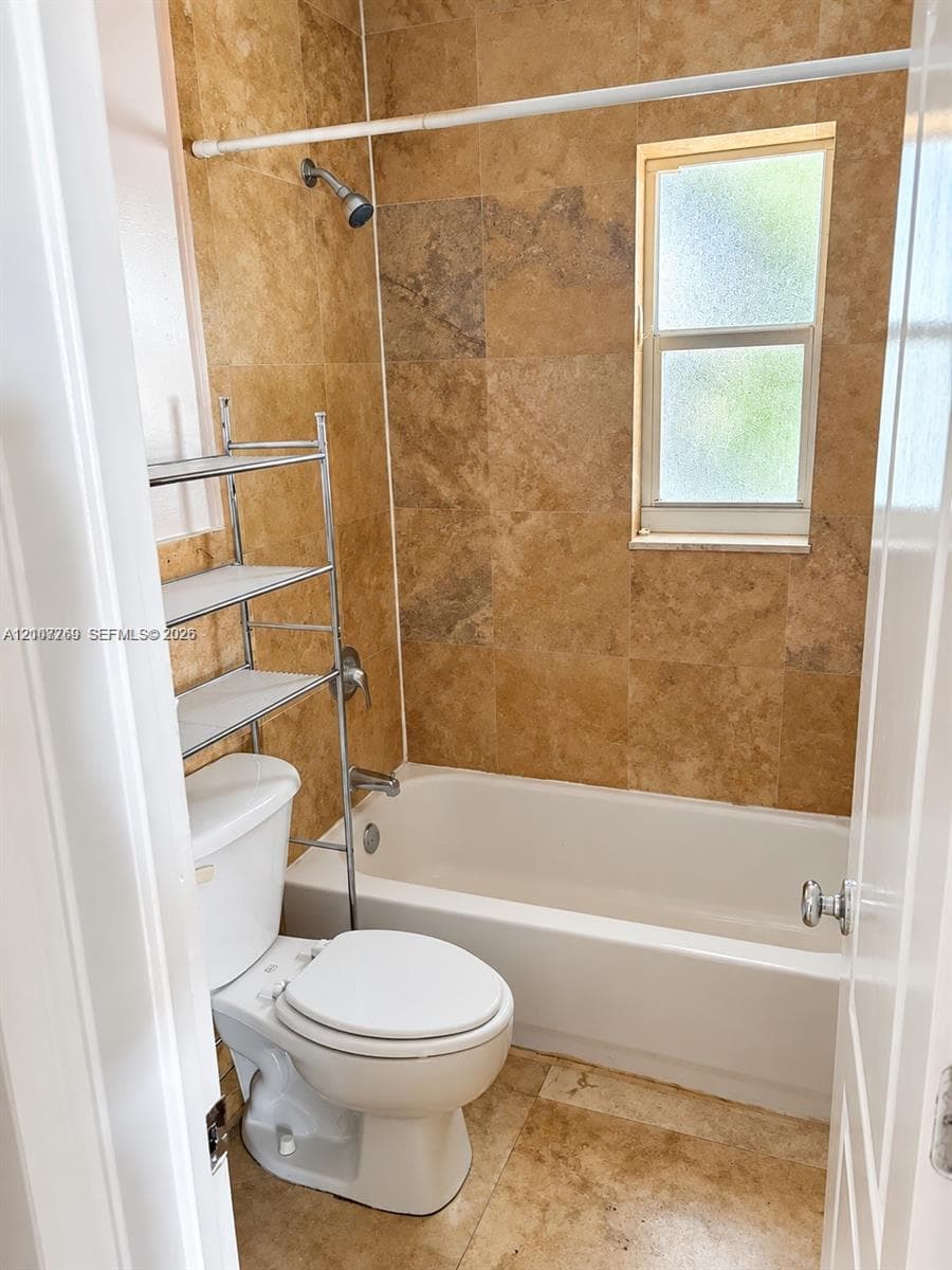 Full-height natural stone wall tile in a warm finish, standard bathtub, and obscured-glass window