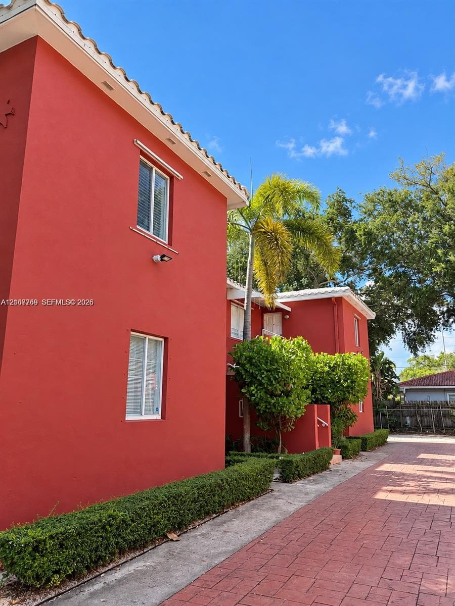 Stucco exterior featuring tile roof accents, multi-pane windows, and a paved brick-style driveway bordered by manicured hedges