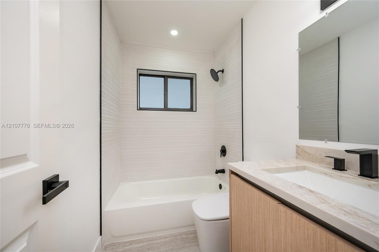 Bathroom featuring wood-finish cabinetry, stone-style countertop with an integrated rectangular sink, matte black fixtures, and textured white tile surround in the tub area