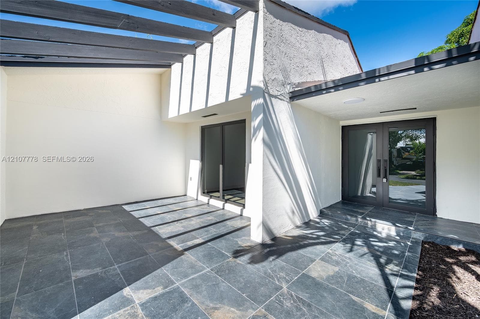 Covered patio featuring charcoal-toned stone flooring, an overhead trellis, double glass-paneled entry doors, and neutral exterior wall finish