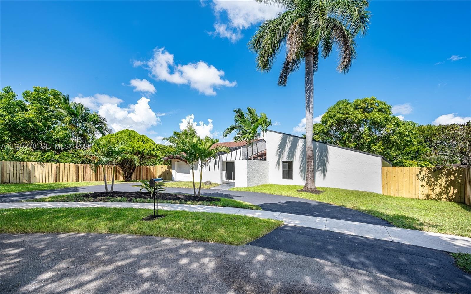 Exterior facade featuring white brick masonry, asphalt driveway, mature palm trees, and perimeter wood fencing