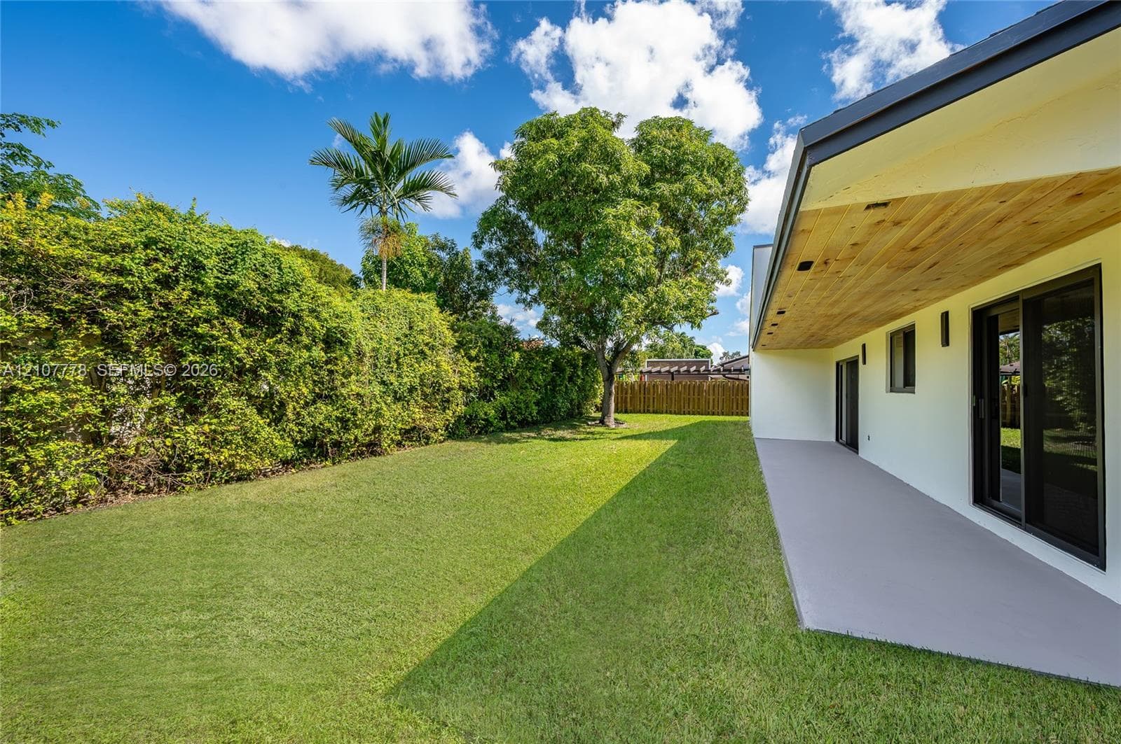 Backyard featuring a concrete patio, wood-paneled ceiling overhang, manicured lawn, and perimeter hedges