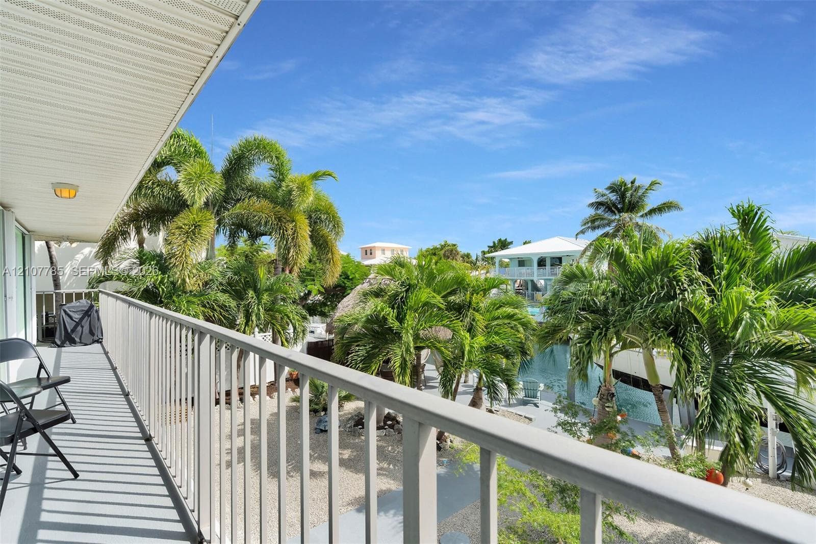 Covered balcony featuring metal railings, overhead lighting, and concrete surfacing, overlooking a canal-front exterior with palm trees and stone ground cover