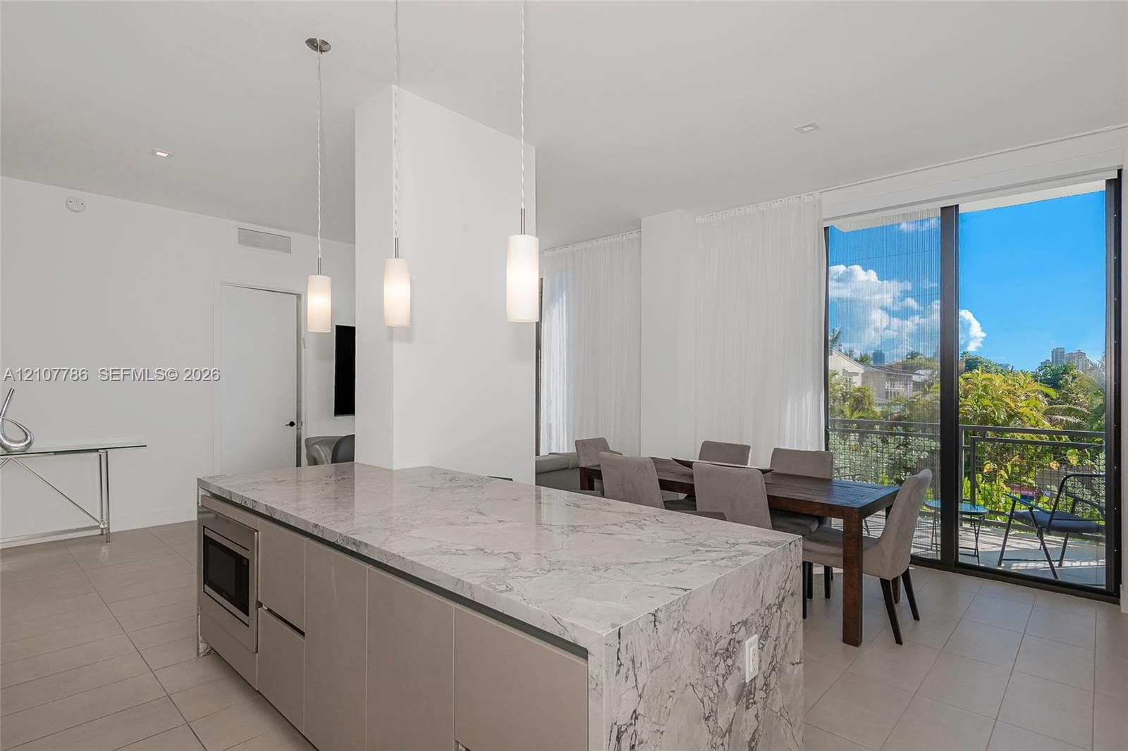Kitchen featuring a waterfall-edge stone island, flat-panel cabinetry, built-in microwave, three pendant lights, and floor-to-ceiling glass doors opening to a balcony