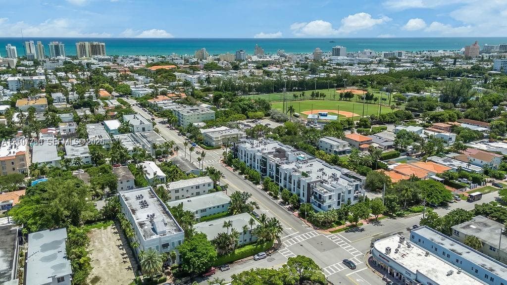 Aerial perspective of an urban neighborhood featuring coastal proximity, multi-story residential structures, mature tree canopy, and proximity to a community ball field