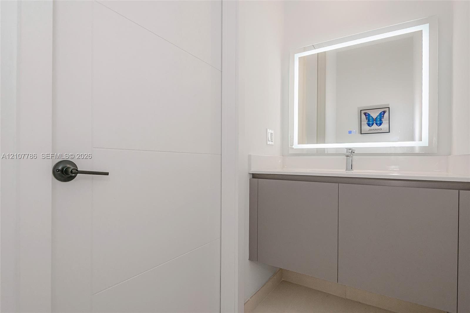 Floating vanity with gray cabinetry and solid-surface countertop, rectangular undermount sink, wall-mounted illuminated mirror, and contemporary matte black lever handle hardware