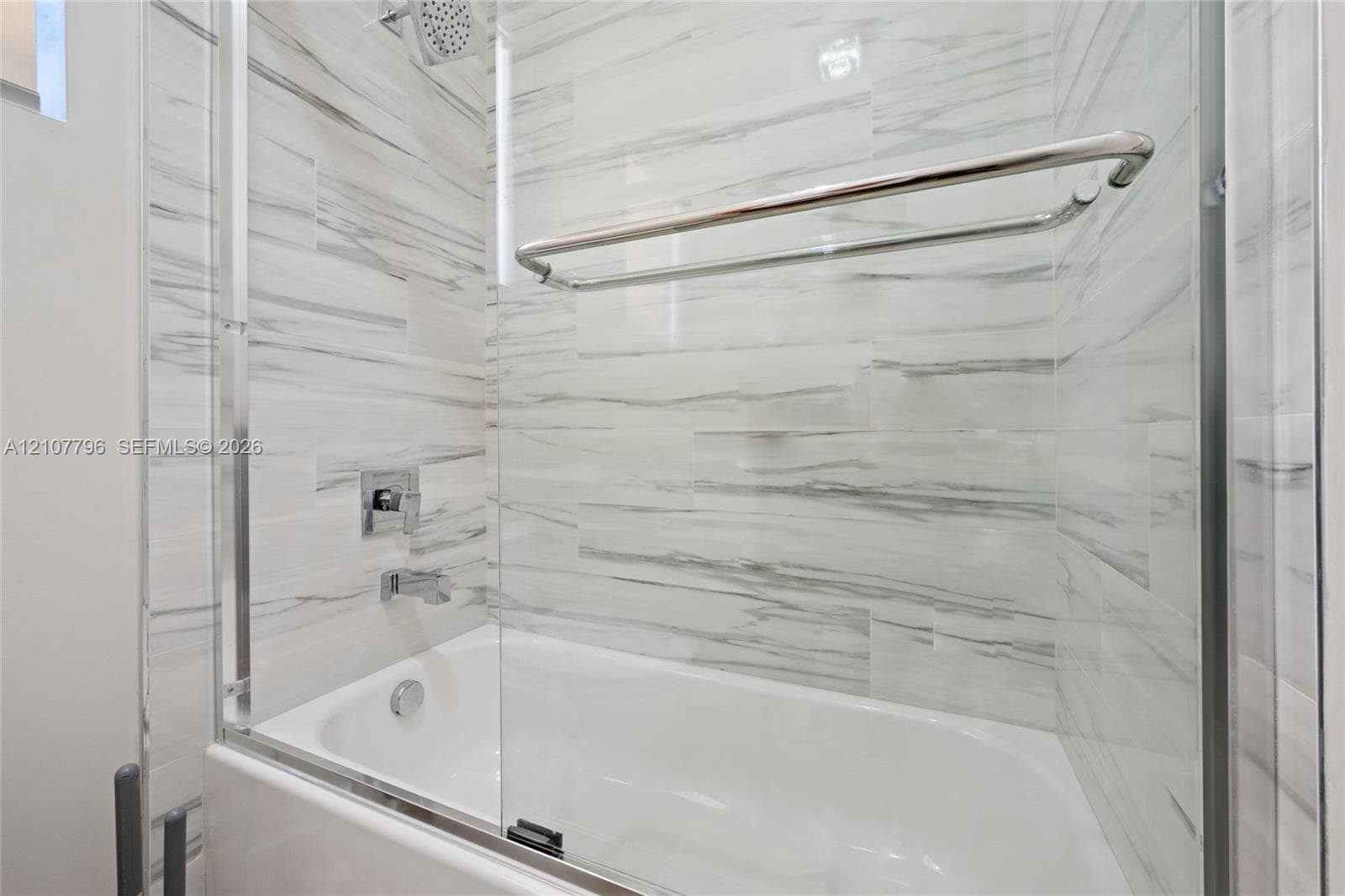 Bathtub featuring floor-to-ceiling stone-look tile surround, glass pivot door, wall-mounted chrome showerhead, and integrated chrome towel bar