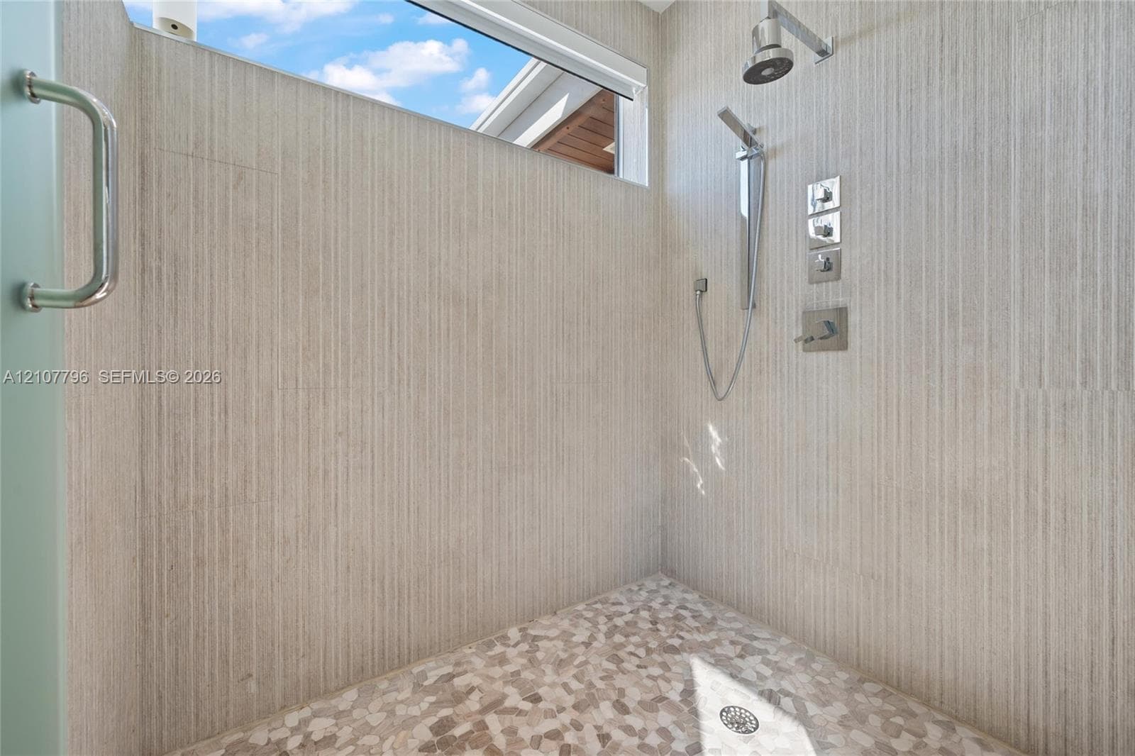 Walk-in shower featuring textured stone-finish wall tile, pebble-style floor tile, a high-mounted rain shower head, a handheld sprayer, and an elongated transom window
