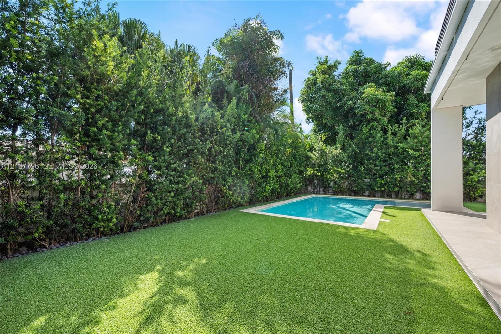 Private backyard featuring an in-ground swimming pool, synthetic turf lawn, stone paver decking, and mature perimeter hedging