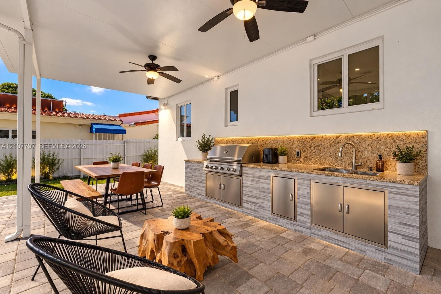 Covered patio featuring paver flooring, two ceiling fans, and an outdoor kitchen with stone veneer base, metal cabinetry, and a stainless steel faucet