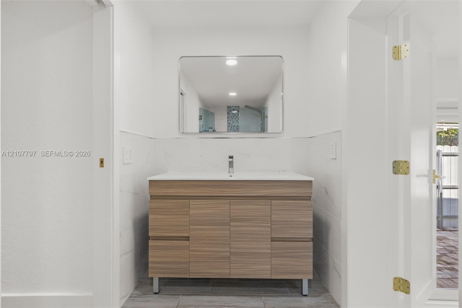Wood-finish vanity with a clean-lined countertop, undermount sink, single-lever chrome faucet, and rectangular mirror with rounded corners, complemented by white stone-finish wall tile