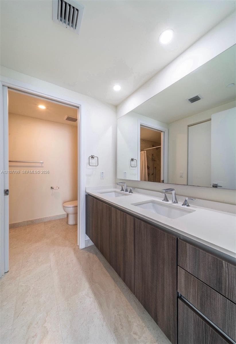 Dual-sink floating vanity with wood-finish cabinetry, solid surface countertop, large wall-mounted mirror, and tile flooring