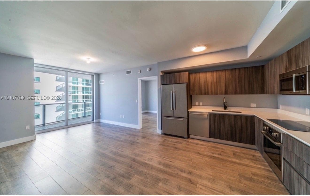 Open-concept kitchen featuring dark wood-finish cabinetry, solid surface countertops, stainless steel appliances, and wood-finish flooring with floor-to-ceiling glass paneling