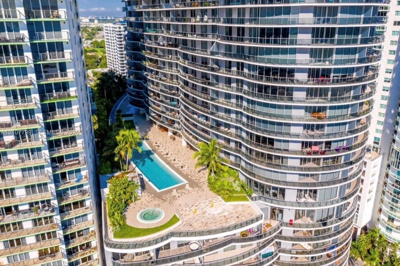 High-rise exterior featuring curved glass balconies, a rectangular swimming pool, a circular spa, tiered hardscaping, and tropical landscaping