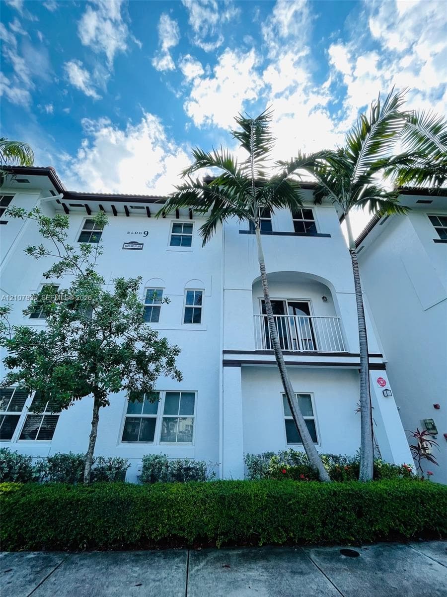 Multi-story building facade featuring white stucco finish, decorative bracket eaves, a private balcony with metal railing, and mature palm landscaping