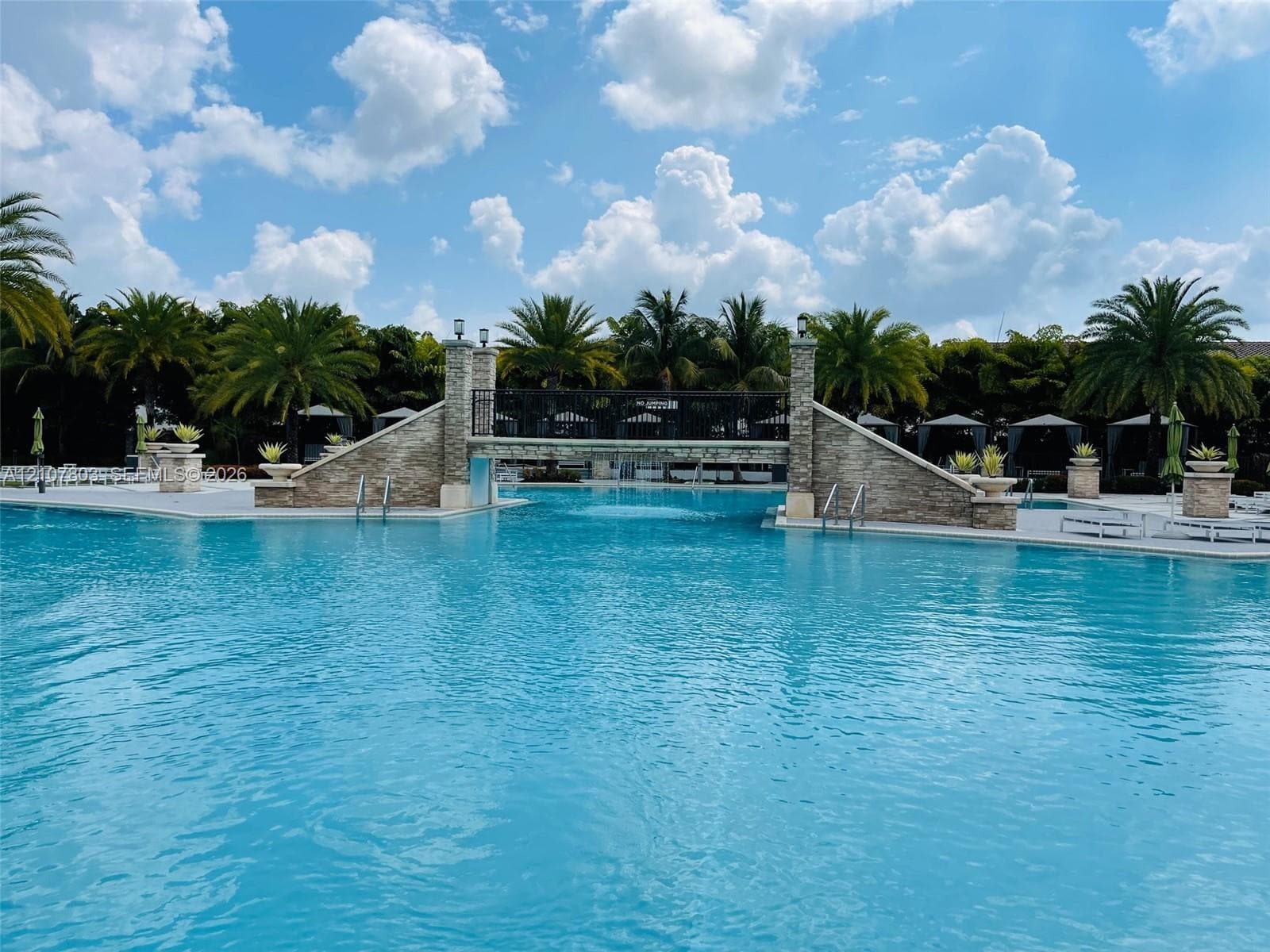 Swimming pool featuring a stone-clad pedestrian bridge, metal railings, integrated stone planters, and paved decking