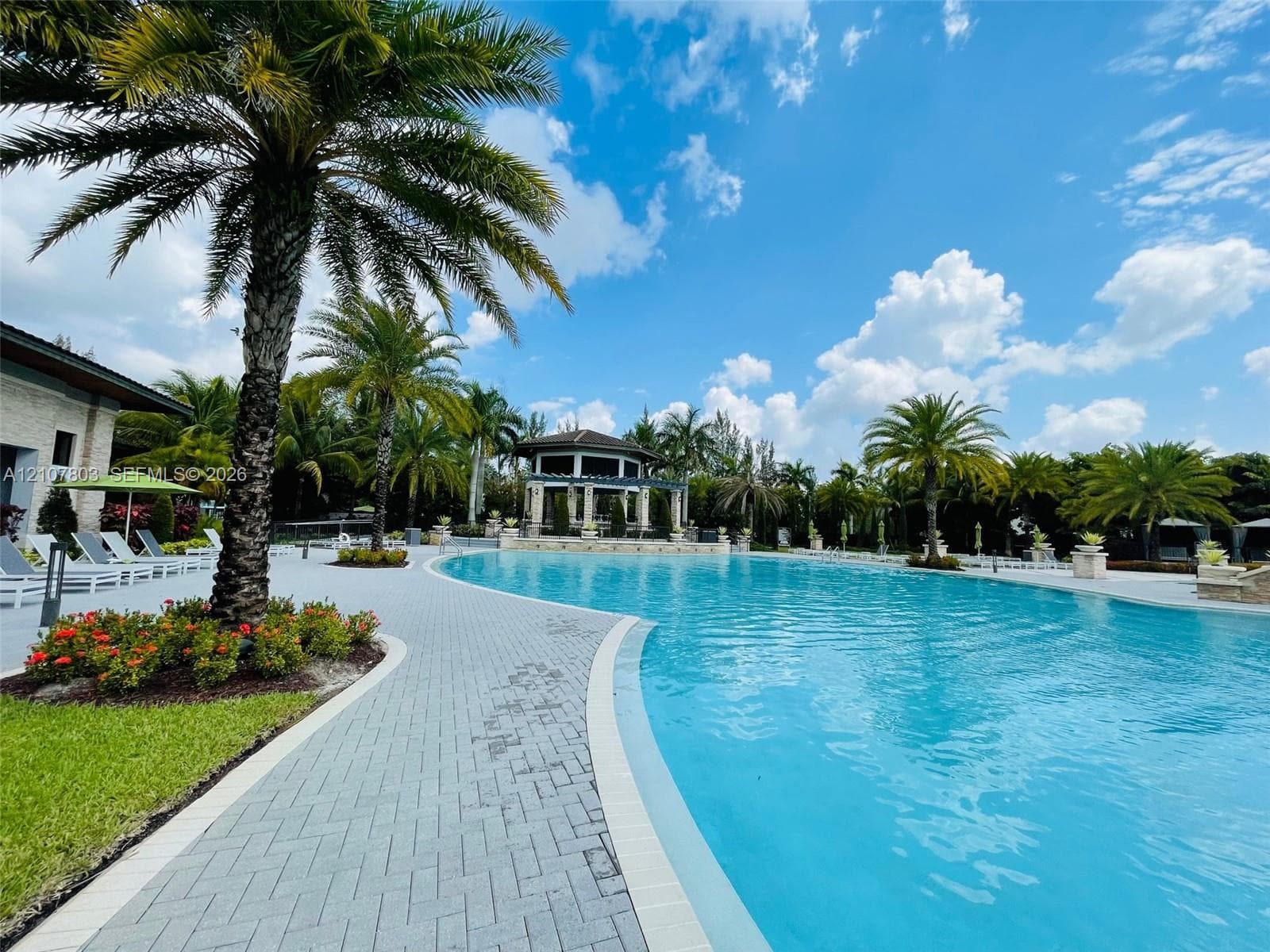 Curved swimming pool featuring a brick-paver deck, integrated gazebo, and mature palm landscaping