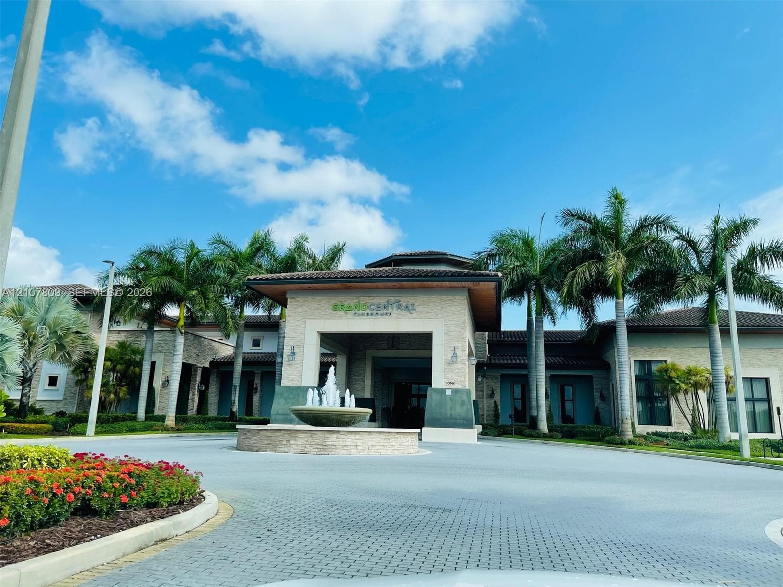 Clubhouse exterior featuring a stone-clad facade, covered porte-cochère, decorative water fountain, and mature palm trees