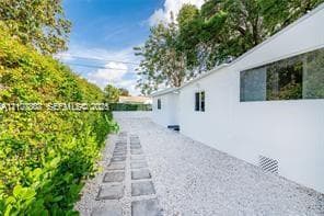 Exterior side yard featuring white stucco siding, stone paver walkway, white gravel landscaping, and established privacy hedges