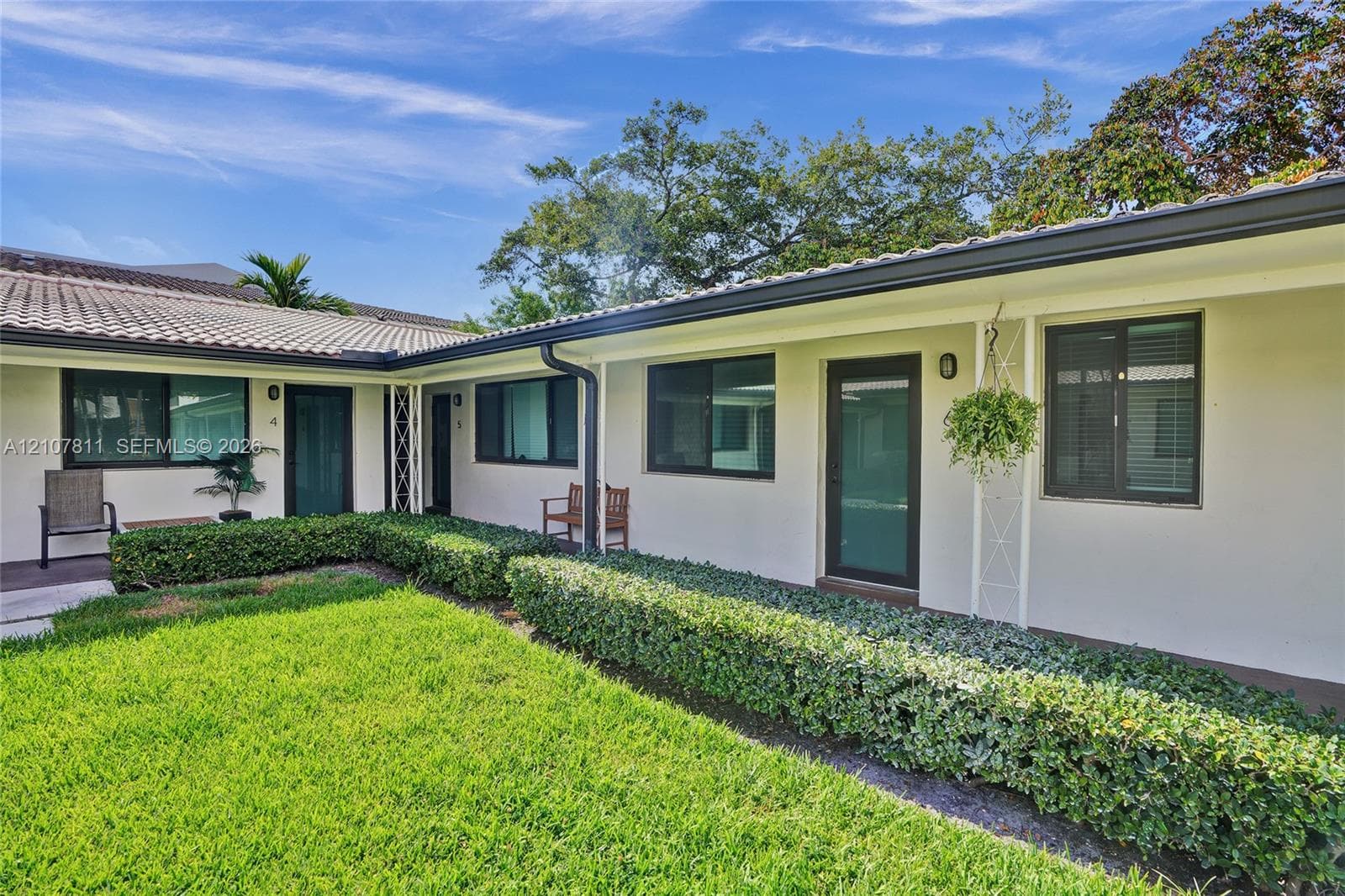 Single-story exterior featuring a light-colored stucco facade, dark-trimmed window frames, tiled roofing, and manicured shrubbery borders