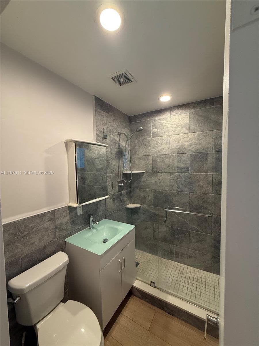 Bathroom featuring a walk-in shower with large-format stone-finish tile surround, glass enclosure, and wall-mounted grab bar, accompanied by a vanity with a glass sink basin and wood-finish flooring