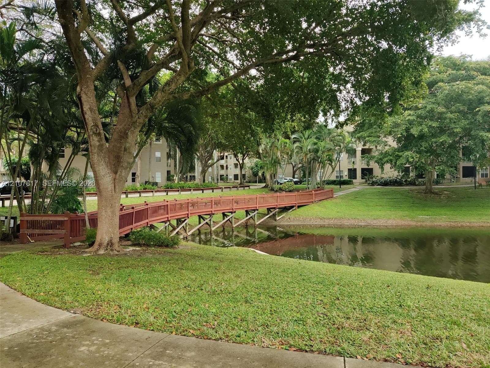 Waterfront setting featuring a wooden pedestrian bridge, manicured landscaping, mature trees, and concrete paved pathways
