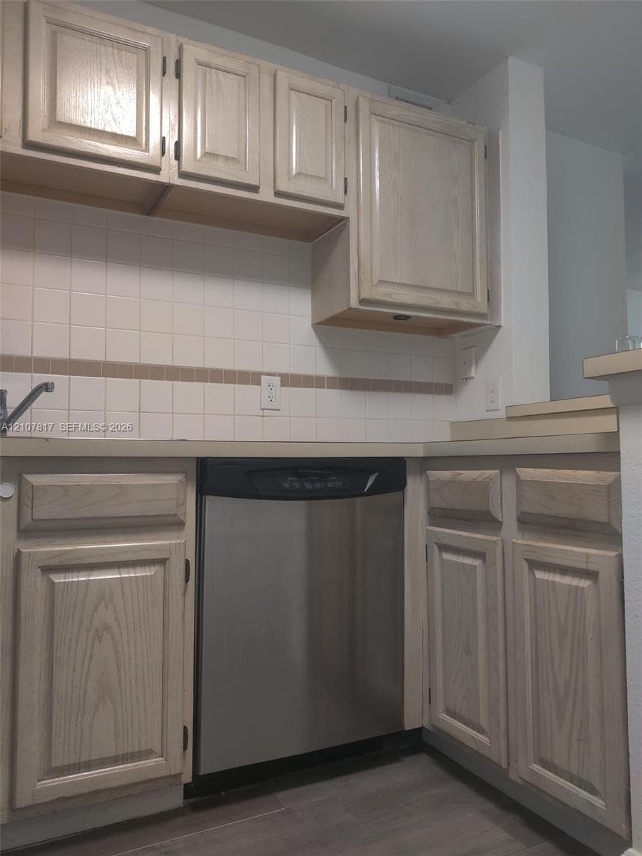 Kitchen featuring light-finish wood cabinetry, stainless steel dishwasher, square tile backsplash with decorative accent inlay, and wood-finish flooring