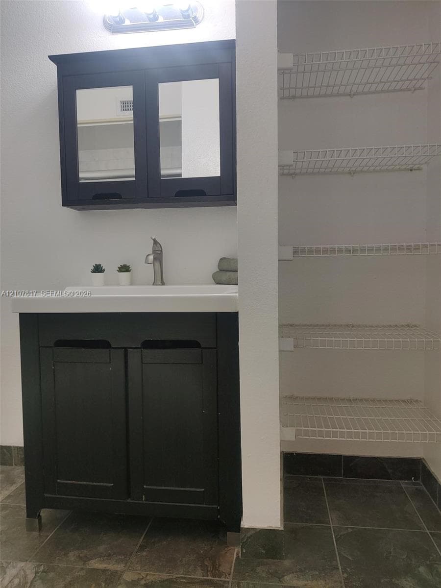 Dark-finish vanity with integrated rectangular sink and matching mirrored cabinet, accompanied by a multi-level wire shelving unit and tile flooring