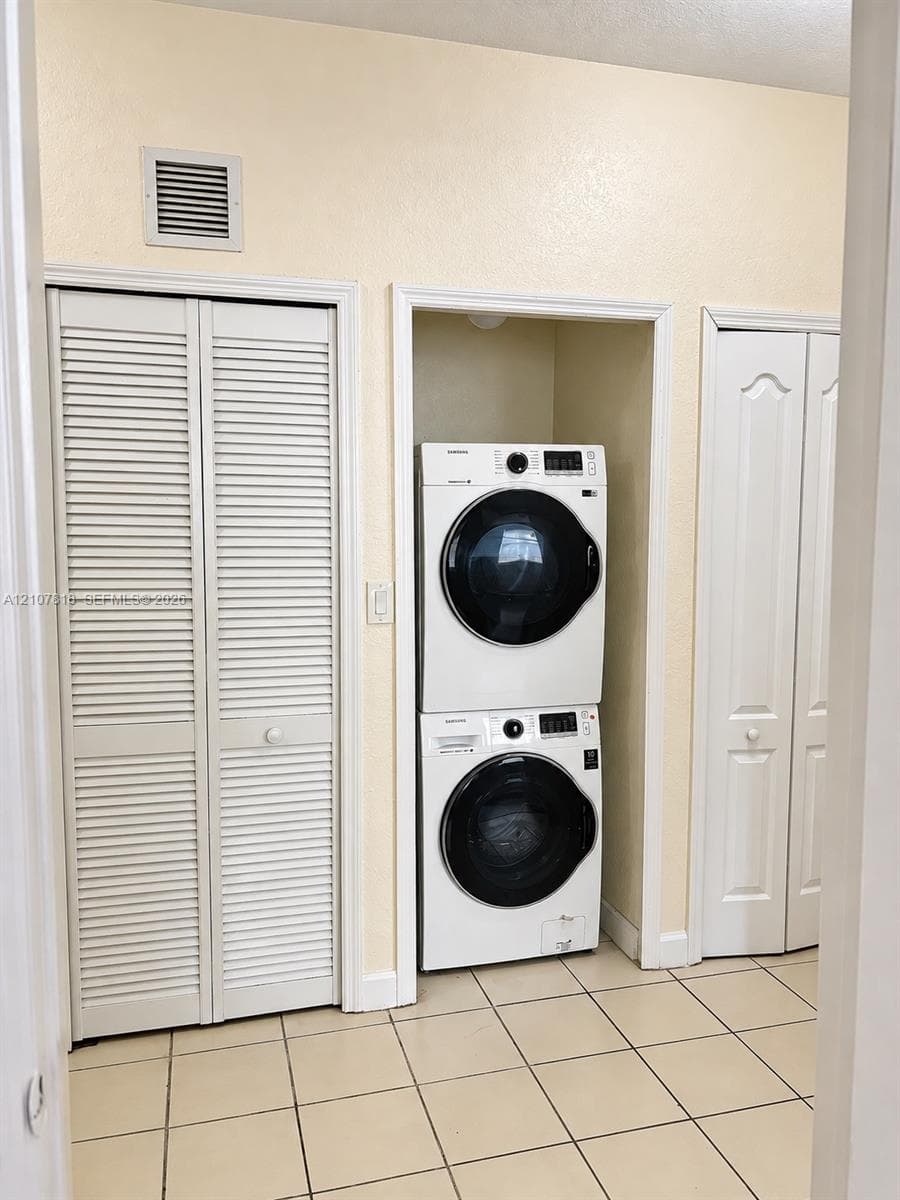 Laundry area featuring a stacked washer and dryer unit, neutral ceramic tile flooring, and dual white louvered closet doors