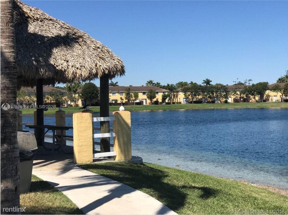Covered thatched-roof pavilion featuring masonry columns with white railing and a concrete walkway situated along the shoreline of a lake