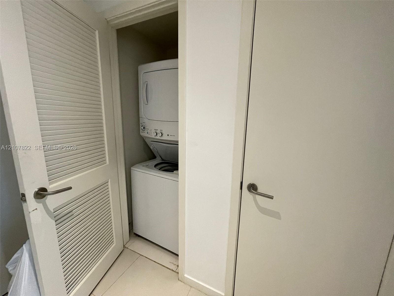 Laundry closet featuring a stacked white appliance unit, louvered swing door, and tile flooring
