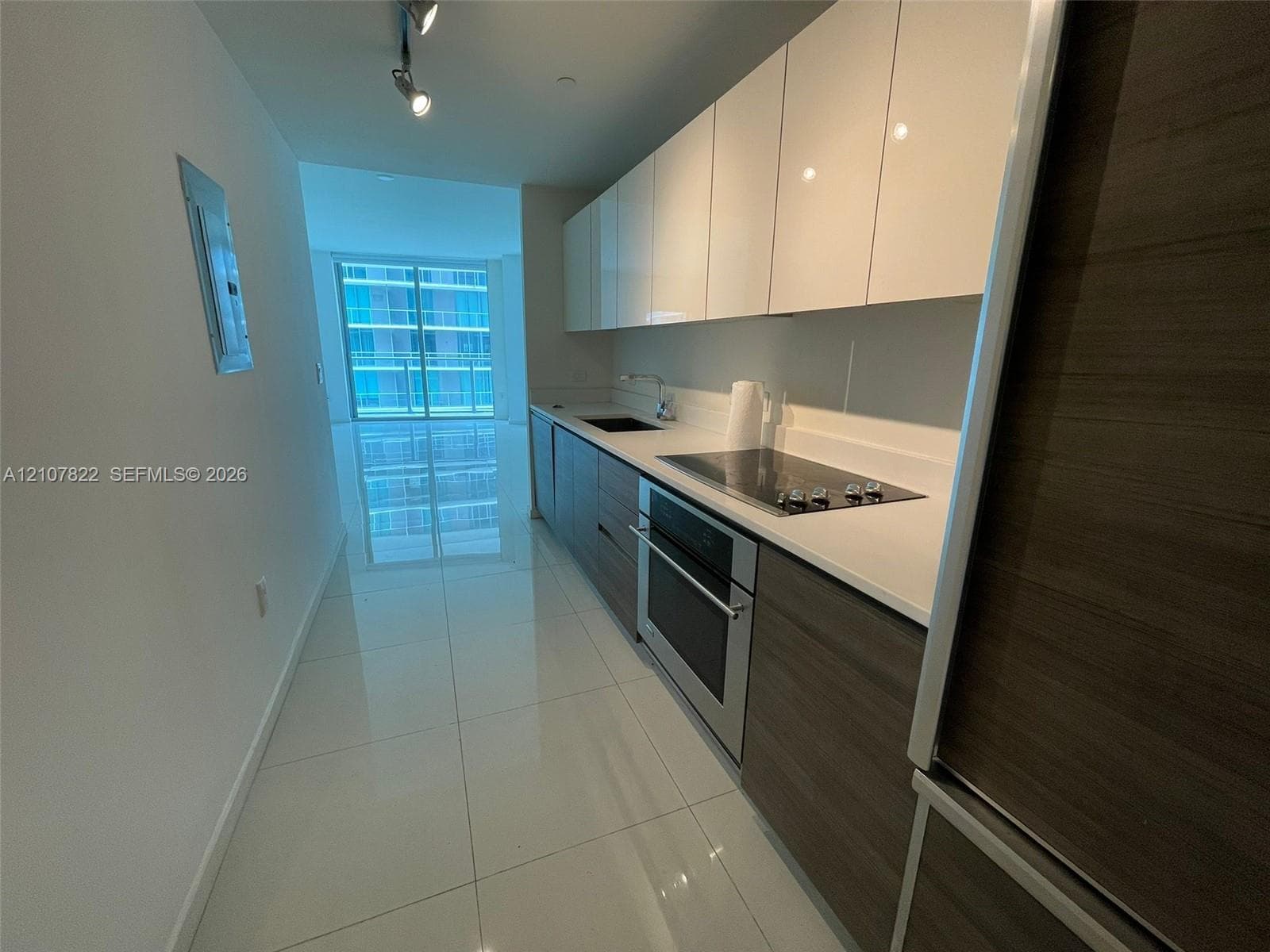 Galley kitchen featuring high-gloss white upper cabinetry, wood-finish lower cabinetry, stone-surface countertops, stainless steel built-in oven, and polished tile flooring