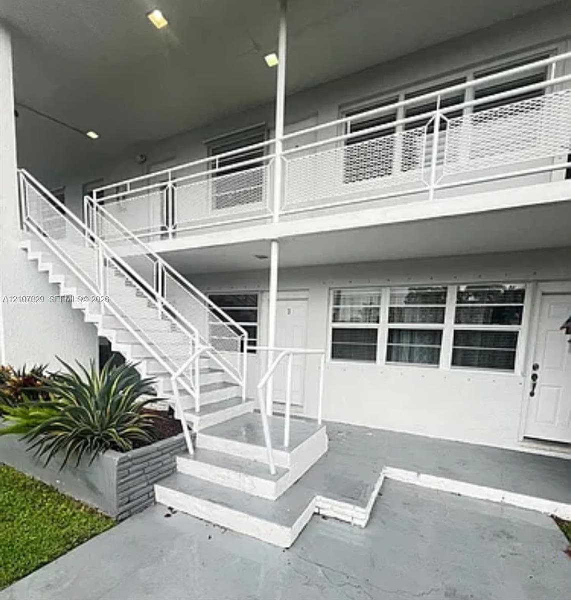Two-story exterior featuring white-painted masonry, metal staircases with diamond-patterned railings, a paved ground-level entry, and a concrete planter box
