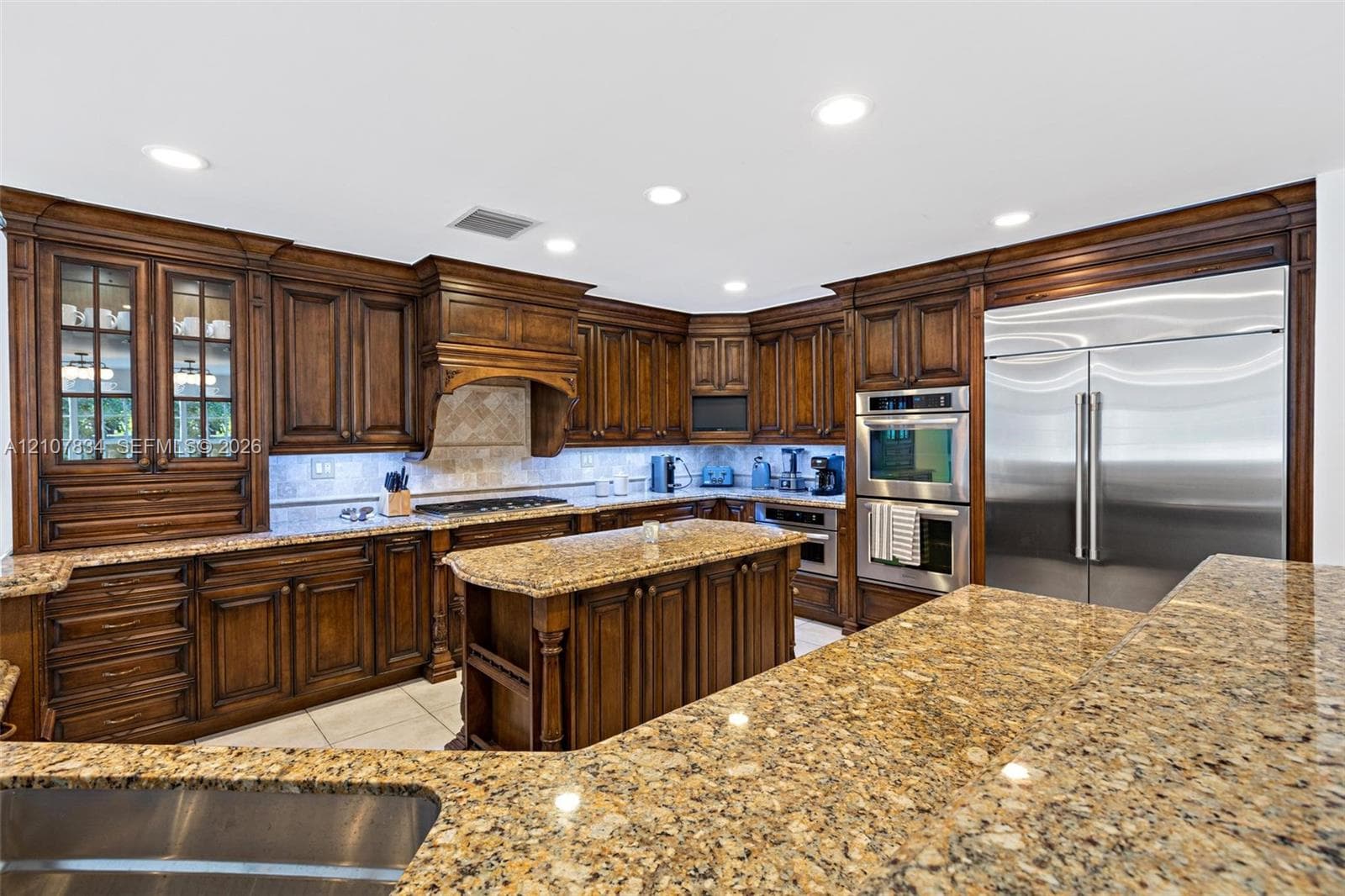 Kitchen featuring custom stained-wood cabinetry with glass-front display panels, natural stone countertops, a central island, recessed lighting, and integrated stainless steel double wall ovens and refrigerator