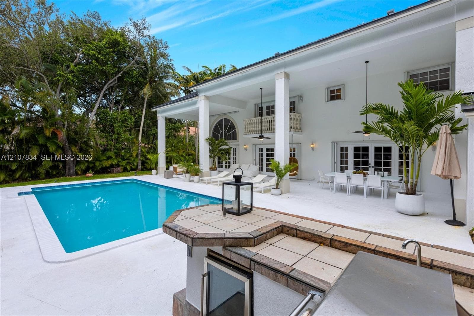 Rear exterior featuring a covered patio, rectangular swimming pool, outdoor kitchen with tiled countertop, second-story balcony, and mature perimeter landscaping