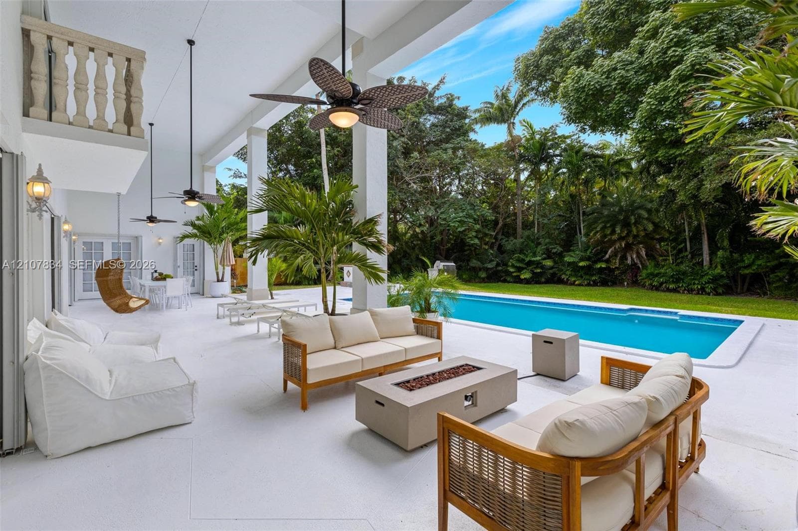 Covered patio featuring white tile flooring, structural columns, ceiling-mounted fans, and a second-story stone balustrade, opening to a rectangular swimming pool and expansive garden area