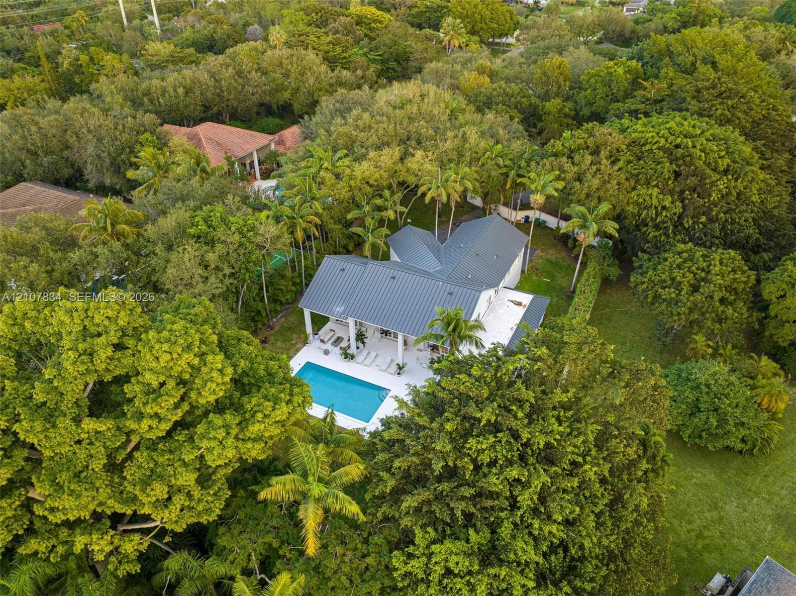 Aerial perspective of a property featuring a metal roof, an in-ground swimming pool, a covered patio with columns, and mature landscaping