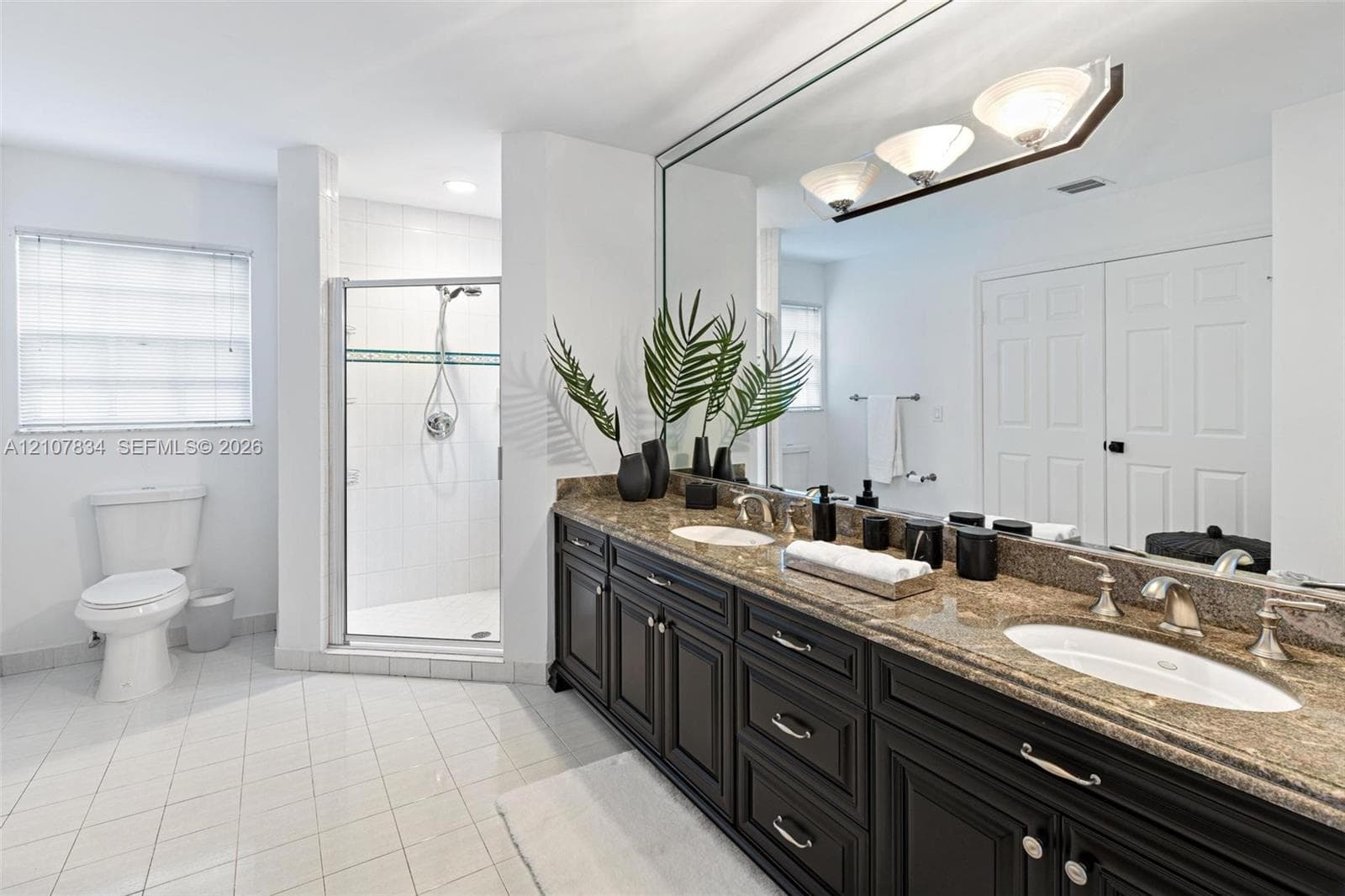 Bathroom featuring dark-finish cabinetry, dual undermount sinks, stone-finish countertops, full-width wall mirror, and glass-enclosed shower