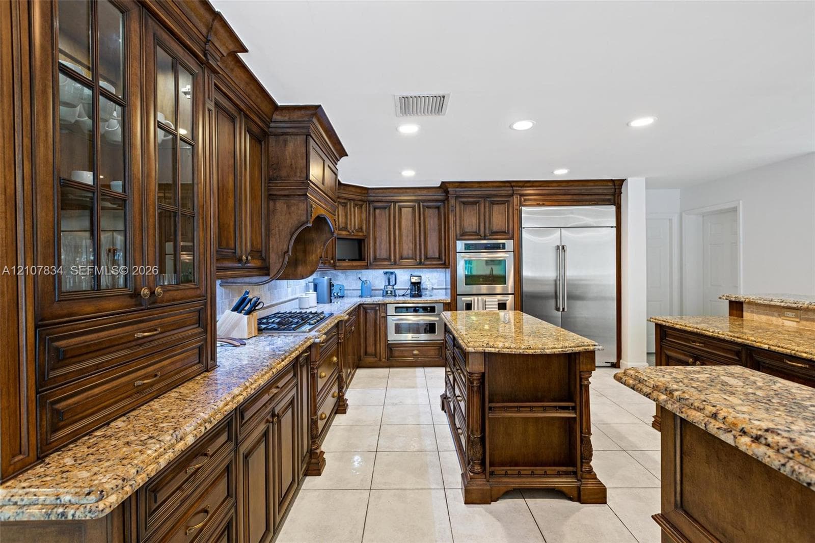 Custom cabinetry with glass-front display panels, stone countertops, central kitchen island, professional-grade stainless steel appliances, and tile flooring