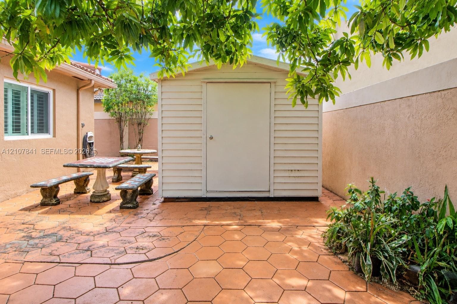 Exterior space featuring a neutral-toned storage shed, hexagonal paver hardscaping, and a stucco-finished wall