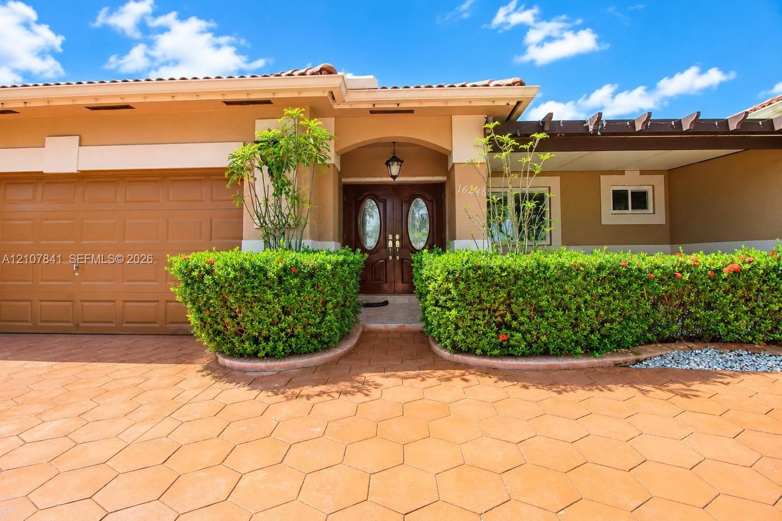 Stucco exterior featuring an arched entryway, double-door entry, attached garage with paneled door, decorative wood-beam pergola, and geometric paver driveway