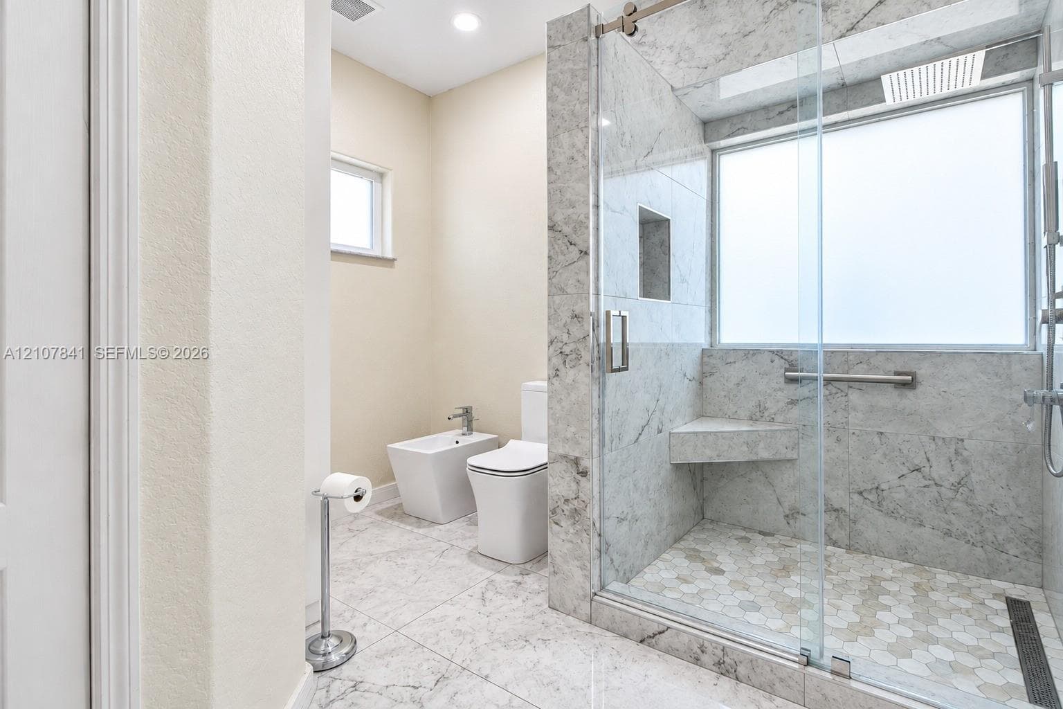 Walk-in shower with floor-to-ceiling stone-finish tile, glass enclosure, built-in bench, and hexagonal floor tile, accompanied by a bidet and dual-flush toilet