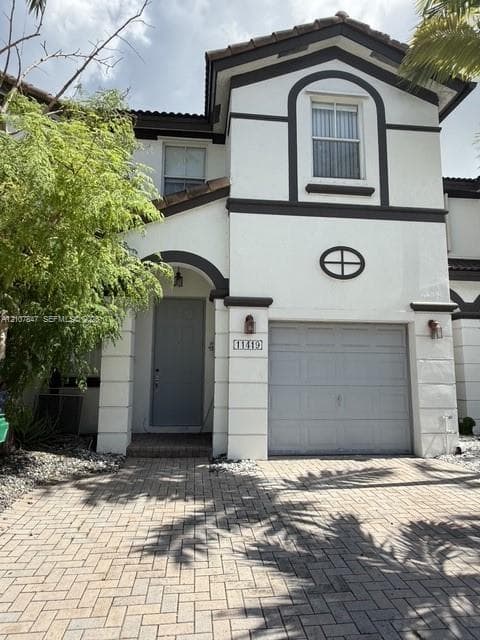 Two-story exterior featuring a stucco facade, arched entryway, brick-patterned paver driveway, single-car garage, and decorative oval window detailing