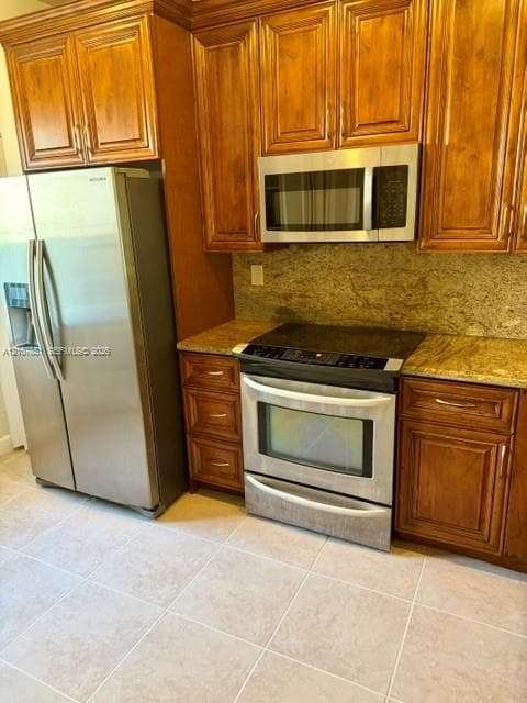 Kitchen featuring stained wood cabinetry, stone countertops, tiled flooring, and stainless steel appliances including a range, microwave, and refrigerator